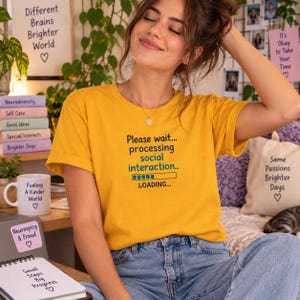 May include: A mustard yellow t-shirt featuring the black and teal text Please wait... processing social interaction... LOADING... with a loading bar graphic. The shirt is paired with blue denim jeans, creating a casual and comfortable look.
