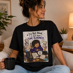 Op de afbeelding: Een zwart T-shirt met korte mouwen en een grafische illustratie van een persoon aan een bureau met de tekst "I'm Not Blind... I Just Have TIME BLINDNESS." Het ontwerp bevat klokken, kalenders, plakbriefjes en een koffiemok.
