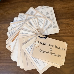 May include: A fan-shaped deck of cream-colored cards bound by a dark blue ring. The top card features the text Cognitive Biases & Logical Fallacies with abstract brown shapes. Each card contains printed text and numbered points regarding psychological concepts.