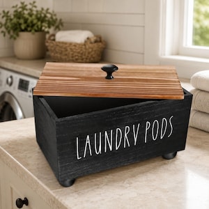 Può includere: Una scatola rettangolare in legno nero con la scritta LAUNDRY PODS stampata in bianco sul davanti. La scatola presenta un coperchio in legno naturale con un pomello nero e poggia su quattro piccoli piedini neri. Il contenitore è aperto in alto.