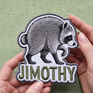 以下が含まれることがあります： 横向きのアライグマの白黒の緻密なイラストが描かれた布製ワッペン。アライグマの下には、オリーブグリーンの太い大文字で「JIMOTHY」という名前が刺繍されています。ワッペンにはダークグレーのステッチ縁取りが施されています。