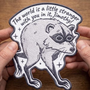 Puede incluir: Un parche bordado con una ilustración de un mapache gris y blanco, el texto "The world is a little stranger with you in it, Jimothy!" y pequeños detalles de estrellas, con un borde cosido en gris oscuro sobre fondo blanco.