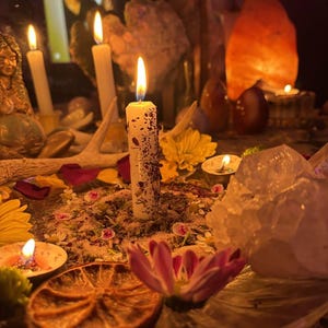 May include: A white pillar candle embedded with dried herbs and flower petals, lit and centered on a surface covered in loose botanical matter. The display includes a dried orange slice, yellow and pink flowers, a large clear quartz crystal, and antlers.