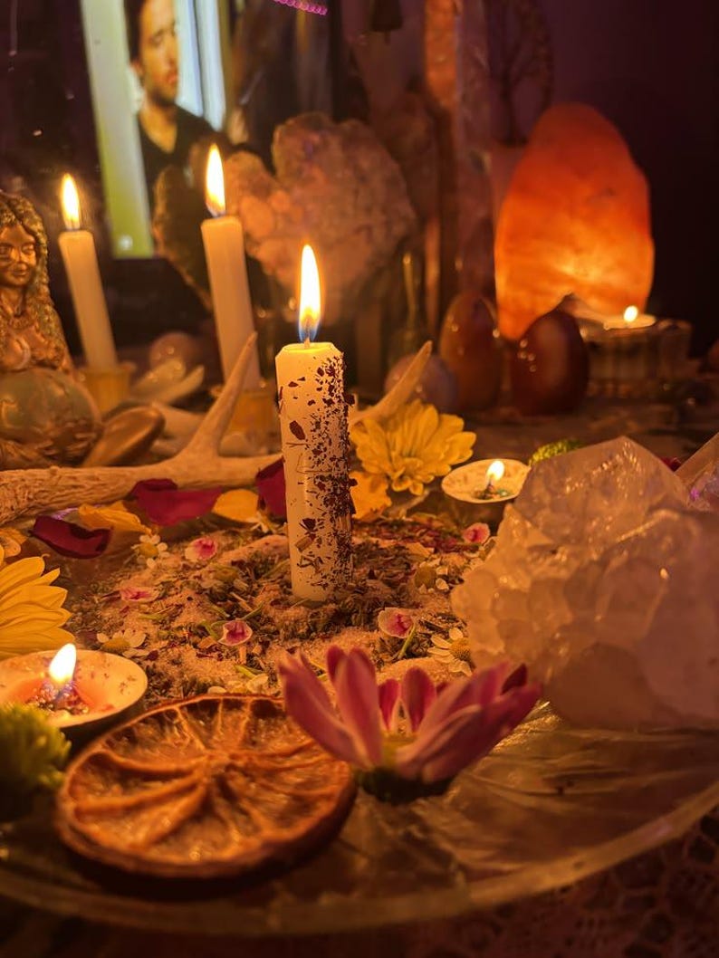 May include: A white pillar candle embedded with dried herbs and flower petals, lit and centered on a surface covered in loose botanical matter. The display includes a dried orange slice, yellow and pink flowers, a large clear quartz crystal, and antlers.