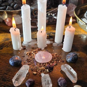 May include: Five white pillar candles with lit flames are arranged in a semi-circle around a central pink rose quartz stone. The stone sits atop a small pile of dried herbs and is surrounded by various polished and raw crystals on a wooden surface.