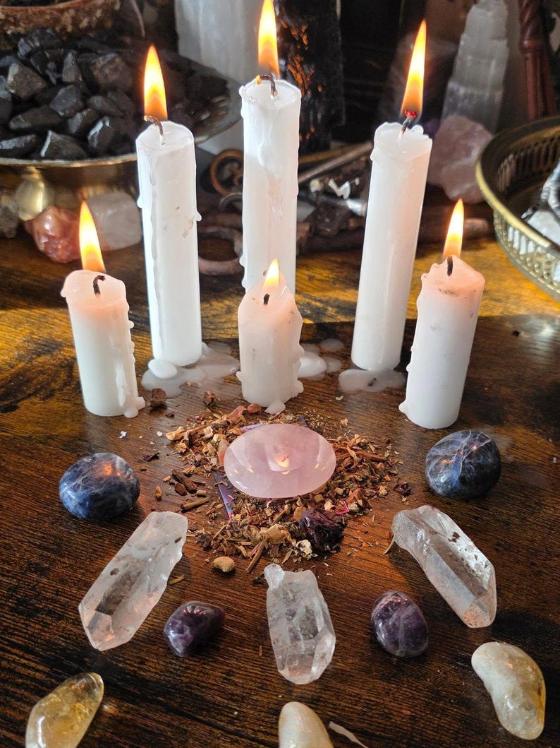 May include: Five white pillar candles with lit flames are arranged in a semi-circle around a central pink rose quartz stone. The stone sits atop a small pile of dried herbs and is surrounded by various polished and raw crystals on a wooden surface.