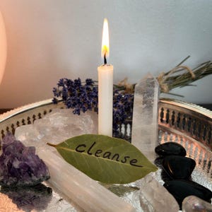 May include: A silver tray holding a lit white taper candle, a green bay leaf with the word cleanse written in black, a purple amethyst cluster, a clear quartz point, a white selenite wand, black stones, and a bundle of dried lavender sprigs.