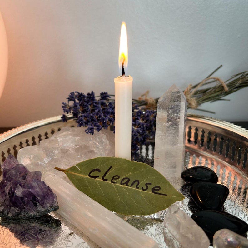 May include: A silver tray holding a lit white taper candle, a green bay leaf with the word cleanse written in black, a purple amethyst cluster, a clear quartz point, a white selenite wand, black stones, and a bundle of dried lavender sprigs.