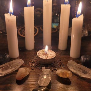 May include: Five tall white pillar candles arranged in a semi-circle around a central tea light candle. The tea light sits on a bed of dried herbs. Several clear and dark polished crystals are placed in a circular formation around the candles.