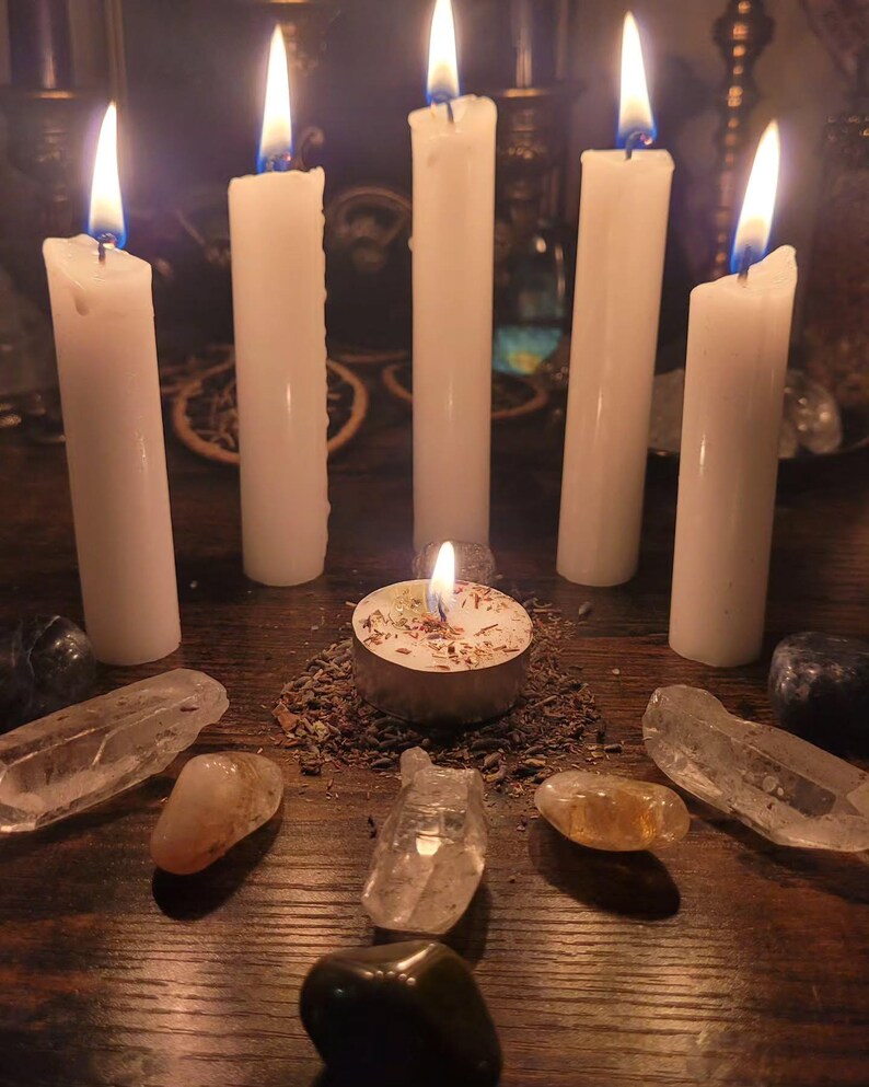 May include: Five tall white pillar candles arranged in a semi-circle around a central tea light candle. The tea light sits on a bed of dried herbs. Several clear and dark polished crystals are placed in a circular formation around the candles.