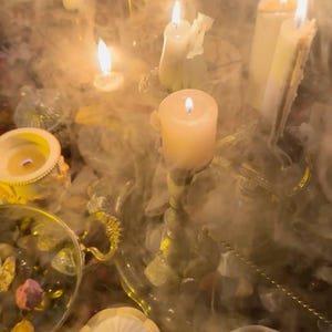 May include: A collection of lit white pillar and taper candles arranged on a gold-toned tray, surrounded by thick swirling smoke. The scene includes small decorative bowls filled with various colorful stones and crystals, all illuminated by the warm glow.