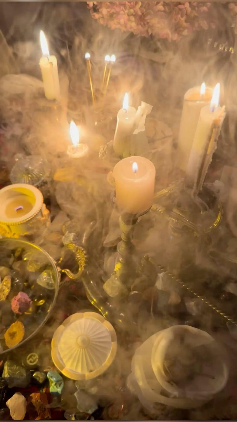 May include: A collection of lit white pillar and taper candles arranged on a gold-toned tray, surrounded by thick swirling smoke. The scene includes small decorative bowls filled with various colorful stones and crystals, all illuminated by the warm glow.