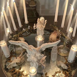 May include: A round glass-topped table featuring a central white angel statue with outstretched wings, surrounded by multiple lit white taper candles in metal holders, white pillar candles in glass jars, and several animal skulls arranged on the surface.