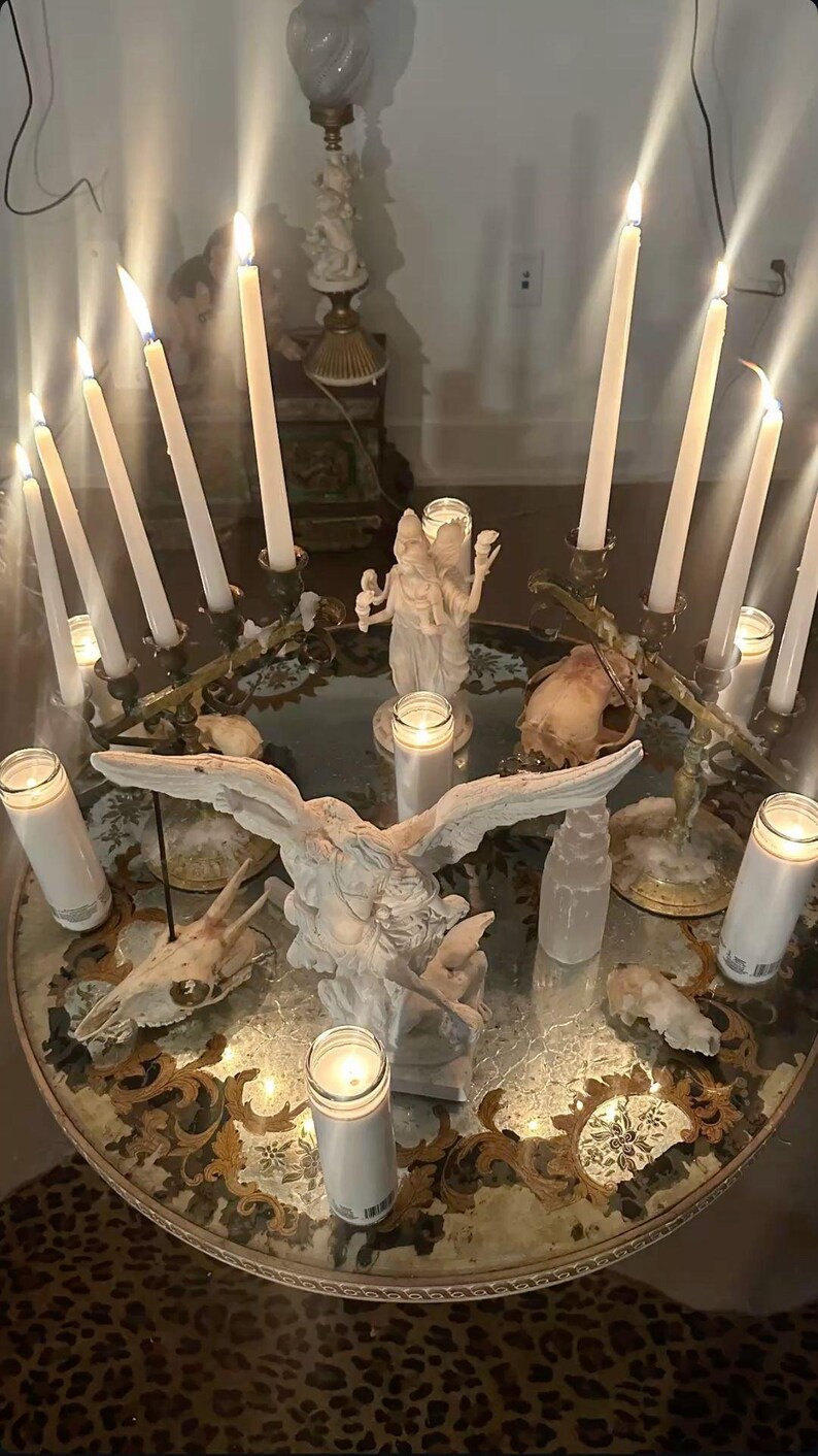 May include: A round glass-topped table featuring a central white angel statue with outstretched wings, surrounded by multiple lit white taper candles in metal holders, white pillar candles in glass jars, and several animal skulls arranged on the surface.