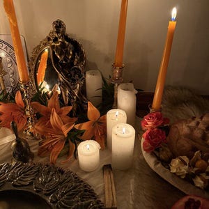 May include: A collection of three tall orange taper candles in ornate gold holders, several white pillar candles, a bronze-toned statue of a figure, orange lilies, red roses, a small bell, a dark textured plate, and a pomegranate on a surface.