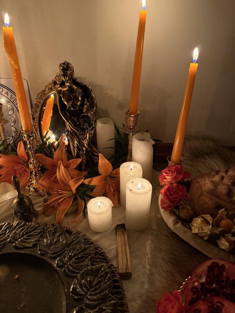 May include: A collection of three tall orange taper candles in ornate gold holders, several white pillar candles, a bronze-toned statue of a figure, orange lilies, red roses, a small bell, a dark textured plate, and a pomegranate on a surface.