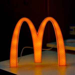 May include: A glowing orange, three-dimensional letter M shaped like the McDonald's golden arches logo. The structure features a visible cross-hatch texture on its surface and is illuminated from within, casting a warm light on the surface below.