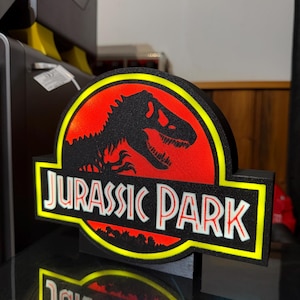 Peut inclure: Une enseigne logo Jurassic Park imprimée en 3D avec une silhouette de T-Rex noire sur un fond circulaire rouge vif, entourée d'une bordure jaune. Le texte JURASSIC PARK est affiché en lettres capitales blanches avec un contour noir sur une base noire.