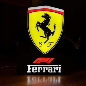 Peut inclure: Une enseigne lumineuse en forme de bouclier arborant le logo jaune Ferrari avec un cheval cabré noir et les lettres S F. Sous le bouclier, une base rectangulaire noire affiche le logo rouge Formule 1 et le texte Ferrari en blanc gras.