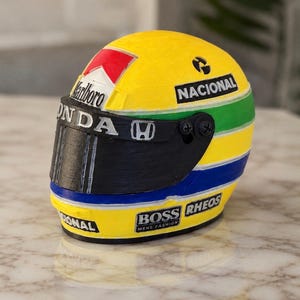 Peut inclure: Un casque de course miniature arborant un motif jaune, vert et bleu. Le casque présente les inscriptions Marlboro, Honda, Nacional, Boss et Rheos. Il est doté d'une visière noire et d'un logo rouge et blanc sur la partie supérieure avant.