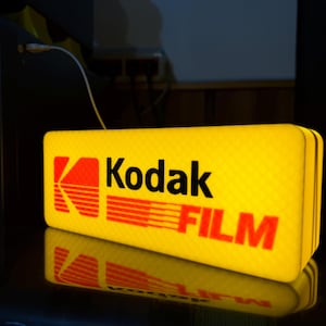 Peut inclure: Une enseigne rectangulaire lumineuse Kodak Film avec un fond jaune vif et un motif quadrillé texturé. Le logo Kodak rouge et jaune est situé à gauche, suivi du texte noir Kodak et du texte rouge FILM.