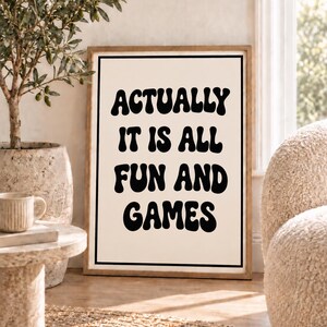 May include: A rectangular framed wall art print featuring a cream-colored background with a thin black border. The text ACTUALLY IT IS ALL FUN AND GAMES is printed in the center in a bold, black, wavy, retro-inspired font style.