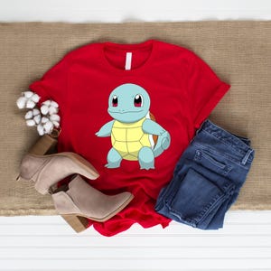 May include: A bright red t-shirt featuring a graphic of a light blue turtle character with a yellow shell and large eyes. The shirt is displayed alongside a pair of blue denim jeans and tan suede ankle boots with a block heel.