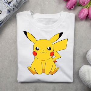 May include: A white short-sleeved t-shirt featuring a centered graphic of the yellow character Pikachu. The character has long ears with black tips, red circular cheeks, and a lightning bolt-shaped tail. The design is printed in vibrant colors.