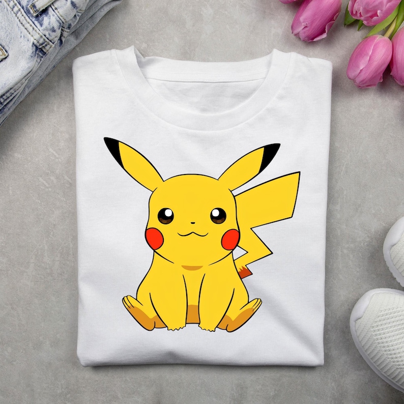 May include: A white short-sleeved t-shirt featuring a centered graphic of the yellow character Pikachu. The character has long ears with black tips, red circular cheeks, and a lightning bolt-shaped tail. The design is printed in vibrant colors.