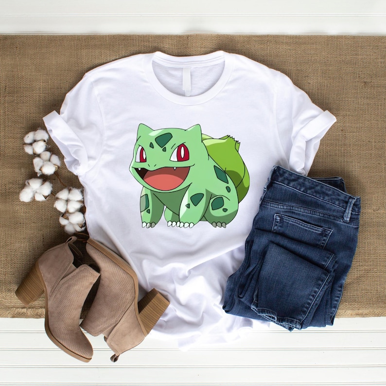 May include: A white t-shirt featuring a graphic of a green creature with red eyes and a large bulb on its back, paired with dark blue denim jeans and tan suede ankle boots with block heels, accented by a sprig of white cotton flowers.