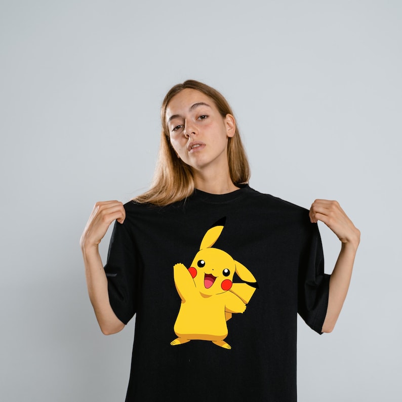 May include: A black short-sleeved t-shirt featuring a centered graphic of the yellow character Pikachu. The character is depicted with its right arm raised, a wide smile, red circular cheeks, and black-tipped ears on a solid black fabric background.