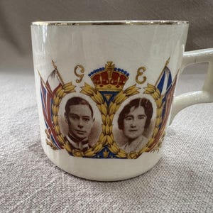 May include: A cream-coloured ceramic mug featuring a gold-rimmed edge and a commemorative graphic. The design displays two sepia-toned portraits of a man and a woman framed by gold laurel wreaths, a central crown, and flanking flags with the letters G and E.