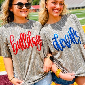 May include: Two short-sleeved crew neck t-shirts with a grey and brown camouflage pattern. One shirt features the word "bulldogs" in a raised, glossy red script font, while the other displays the word "devils" in a raised, glossy blue script font.