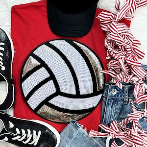 May include: A red t-shirt featuring a large circular volleyball graphic made of white and gold sequins with black outlines. The shirt is paired with a black baseball cap, black sneakers with white laces, blue denim shorts, and a red and white striped ribbon.