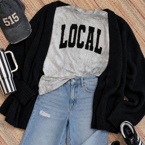 May include: A gray acid-wash t-shirt with the word LOCAL in black block letters, a black open-front cardigan, light blue distressed denim jeans, a dark gray baseball cap with the number 515, and a black and white striped tumbler cup.