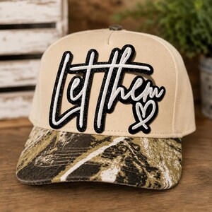 May include: A beige baseball cap featuring a camouflage pattern on the brim. The front panel displays a black and white patch with the script text Let Them and a small heart symbol. The cap has a structured crown and a button on the top center.
