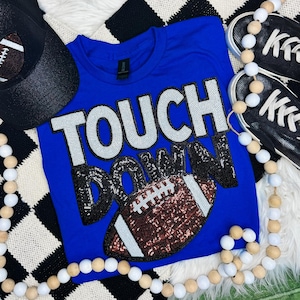 May include: A royal blue t-shirt featuring the word TOUCH in white sequins and the word DOWN in black sequins. Below the text is a brown sequined football graphic with white sequin laces and stripes. A black cap and black sneakers are visible.