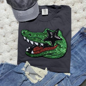 May include: A charcoal gray t-shirt featuring a large, green sequined alligator head patch with a black sequined star over the eye, white teeth, and a red tongue. The shirt is paired with a distressed gray baseball cap and blue denim jeans.