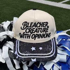 May include: A cream-colored baseball cap featuring the black embroidered text "BLEACHER CREATURE WITH OPINIONS" on the front crown. The brim is black with a white star pattern and a double-line white border detail along the edge.