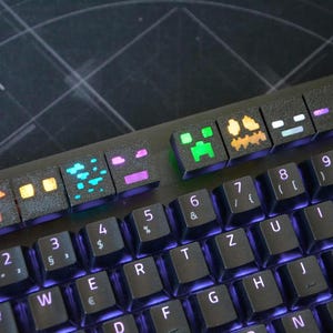 Puede incluir: Un teclado de computadora negro con una fila de teclas personalizadas texturizadas sobre las teclas numéricas. Estas teclas muestran varios iconos pixelados en colores vibrantes, incluyendo naranja, azul, morado y verde, sobre un fondo oscuro.