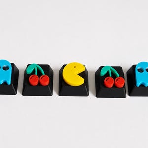 Può includere: Un set di cinque tasti neri per tastiera meccanica. I due esterni presentano figure di fantasmi azzurri, il secondo e il quarto mostrano ciliegie rosse con gambi verdi, e il tasto centrale presenta un personaggio circolare di colore giallo.