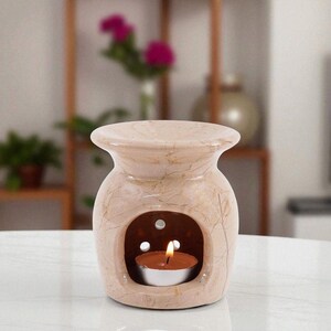 May include: A beige ceramic oil burner with a marbled pattern and a wide, flared top bowl. The base features a large arched opening containing a small, lit tea light candle in a metal cup, with three circular ventilation holes above it.