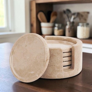 May include: A set of circular beige marble coasters stacked inside a matching cylindrical marble holder. The stone features natural veining patterns in shades of tan and cream. One coaster is positioned upright in front of the holder.