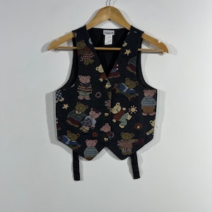May include: A black sleeveless vest featuring a repeating woven pattern of teddy bears wearing sweaters, small stars, and suns. The vest has a V-neckline, gold-toned heart-shaped buttons, and two black fabric straps hanging from the bottom hem.