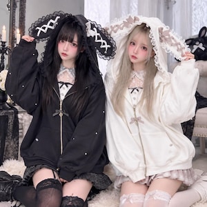 May include: Two zip-up hoodies featuring large rabbit ears with lace trim and ribbon lacing. One hoodie is black with white pearls, and the other is white with beige pearls. Both include a front zip with a small bow and heart-shaped charm.