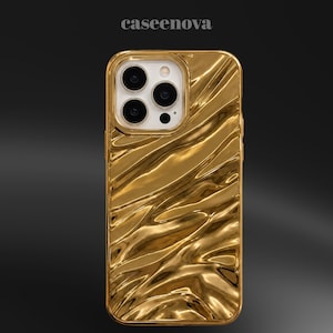 Puede incluir: Una funda para smartphone de color dorado con una textura reflectante, ondulada y similar a un líquido. La funda presenta un recorte para un sistema de cámara de triple lente y el texto caseenova está impreso en letras blancas pequeñas.