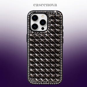 Puede incluir: Una funda de smartphone negra con una cuadrícula de tachuelas piramidales de metal oscuro en la parte posterior, bordeada por un marco de pequeños cristales transparentes. El texto caseenova está impreso en blanco arriba sobre la funda.