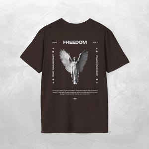 May include: A dark brown t-shirt featuring a white graphic of a winged figure centered on the back. The text reads FREEDOM, 2024, VOL 1, TRUST YOUR INSTINCT, FOLLOW YOUR HEART, and a paragraph about trusting your heart and ideas in white print.
