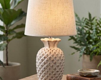 May include: A table lamp featuring a white, textured pineapple-shaped base with a distressed finish and a tapered, off-white fabric lampshade. The lamp sits on a wooden surface next to a small ceramic dish and a vintage book.
