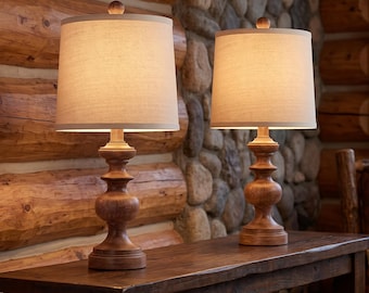 May include: A pair of table lamps featuring turned wooden bases with a warm brown finish and cylindrical off-white fabric shades. The lamps are illuminated, casting a soft glow, and are positioned side by side on a dark wooden surface.